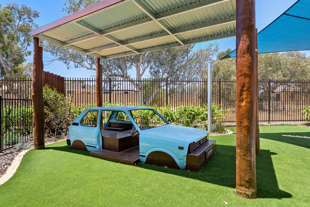 A blue car is situated under a metal canopy on a lawn at Nido Piara Waters child care centre.